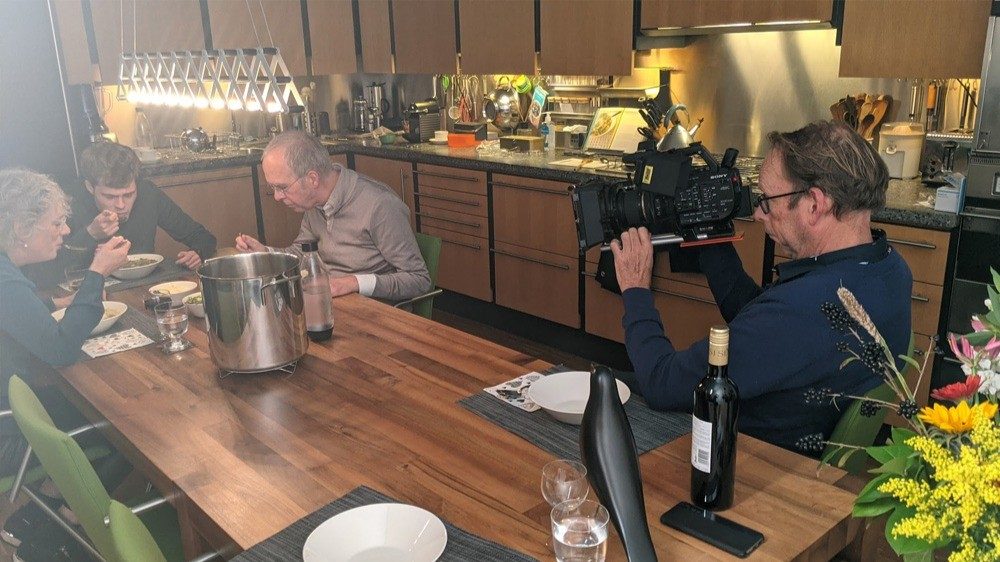Een cameraman filmt een gesprek aan een keukentafel waar mensen soep eten
